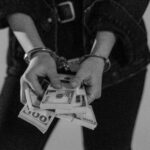 Black and white image of a handcuffed person holding bundles of US dollars.