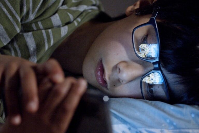 A child lying in bed at night using a smartphone, with screen reflection on eyeglasses.